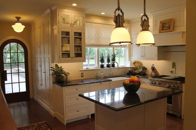 Example of a mid-sized classic u-shaped dark wood floor enclosed kitchen design in Other with a farmhouse sink, shaker cabinets, white cabinets, granite countertops, white backsplash, subway tile backsplash, stainless steel appliances and an island