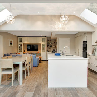 Traditional galley open plan kitchen in London with a submerged sink, shaker cabinets, white cabinets, grey splashback, stone slab splashback, black appliances, an island, beige floors and white worktops.