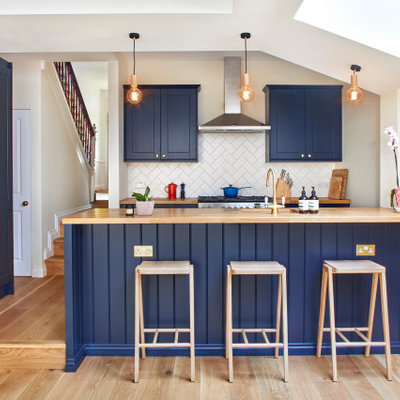 Inspiration for a transitional u-shaped medium tone wood floor and brown floor kitchen remodel in London with an undermount sink, shaker cabinets, blue cabinets, wood countertops, white backsplash, paneled appliances, a peninsula and brown countertops