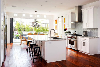 Design ideas for an expansive eclectic galley kitchen/diner in Los Angeles with a built-in sink, shaker cabinets, white cabinets, engineered stone countertops, grey splashback, glass tiled splashback, stainless steel appliances, medium hardwood flooring, an island, brown floors and multicoloured worktops.