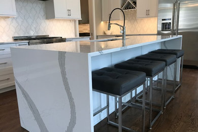 Inspiration for a large modern l-shaped kitchen/diner in New York with shaker cabinets, white cabinets, an island, a submerged sink, quartz worktops, white splashback, stainless steel appliances, dark hardwood flooring and brown floors.