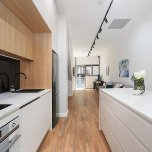 This is an example of a contemporary galley kitchen in Sydney with a drop-in sink, flat-panel cabinets, white cabinets, black splashback, glass sheet splashback, black appliances, medium hardwood floors, with island, brown floor and white benchtop.