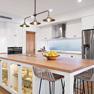 Design ideas for a mid-sized beach style l-shaped open plan kitchen in Perth with a single-bowl sink, shaker cabinets, white cabinets, wood benchtops, white splashback, subway tile splashback, stainless steel appliances, medium hardwood floors, with island, brown floor and brown benchtop.