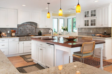 Large trendy u-shaped medium tone wood floor and brown floor eat-in kitchen photo with a farmhouse sink, raised-panel cabinets, white cabinets, granite countertops, gray backsplash, stone tile backsplash, stainless steel appliances and an island