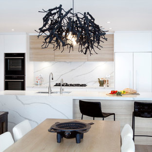 Large contemporary galley open plan kitchen in Sydney with an undermount sink, flat-panel cabinets, white cabinets, quartz benchtops, white splashback, stone slab splashback, black appliances, light hardwood floors, with island and white benchtop.