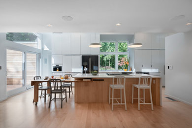 Design ideas for a medium sized contemporary galley open plan kitchen in Boston with a single-bowl sink, flat-panel cabinets, white cabinets, engineered stone countertops, glass sheet splashback, black appliances, light hardwood flooring, an island, white worktops and beige floors.