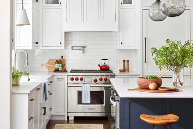 Beach style l-shaped dark wood floor kitchen photo in Boston with shaker cabinets, white cabinets, white backsplash, a farmhouse sink, subway tile backsplash, stainless steel appliances, an island and white countertops