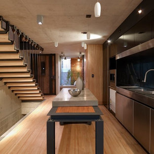 Photo of a mid-sized industrial galley open plan kitchen in Sydney with an integrated sink, flat-panel cabinets, stainless steel cabinets, concrete benchtops, black splashback, glass sheet splashback, stainless steel appliances, light hardwood floors, with island, beige floor and grey benchtop.