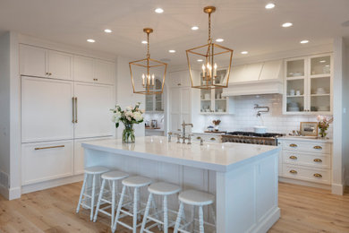 Example of a large transitional medium tone wood floor and brown floor kitchen design in Vancouver with shaker cabinets, white cabinets, white backsplash, ceramic backsplash, paneled appliances, an island, white countertops and a farmhouse sink