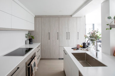 Eat-in kitchen - mid-sized modern l-shaped light wood floor and beige floor eat-in kitchen idea in Vancouver with a single-bowl sink, flat-panel cabinets, beige cabinets, solid surface countertops, white backsplash, stone slab backsplash, stainless steel appliances, an island and white countertops