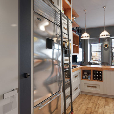 Example of a small trendy galley light wood floor open concept kitchen design in New York with shaker cabinets, a peninsula, a farmhouse sink, white cabinets, wood countertops, white backsplash, ceramic backsplash and stainless steel appliances