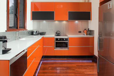 Esempio di una cucina contemporanea di medie dimensioni con lavello integrato, ante lisce, ante arancioni, top in superficie solida, paraspruzzi a effetto metallico, paraspruzzi con lastra di vetro, elettrodomestici in acciaio inossidabile, parquet scuro, nessuna isola e top grigio