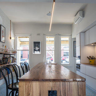 This is an example of an industrial galley kitchen in Melbourne with flat-panel cabinets, white cabinets, wood benchtops, white splashback, brick splashback, stainless steel appliances, with island, multi-coloured floor and brown benchtop.