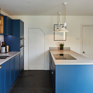 Inspiration for a transitional galley kitchen in London with a double-bowl sink, shaker cabinets, blue cabinets, grey splashback, a peninsula, grey floor, slate floors and white benchtop.