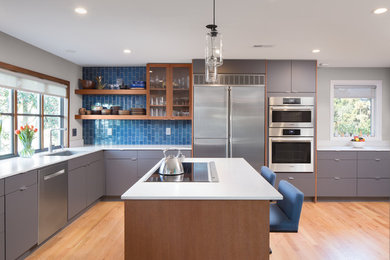 Diseño de cocina actual con armarios con paneles lisos, puertas de armario grises, salpicadero azul, electrodomésticos de acero inoxidable, una isla, encimeras blancas y suelo de madera clara