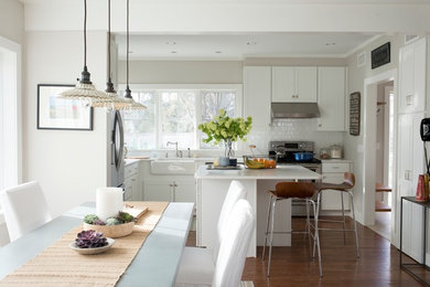 Ejemplo de cocina comedor lineal campestre de tamaño medio con fregadero bajoencimera, armarios estilo shaker, puertas de armario blancas, encimera de cuarzo compacto, salpicadero blanco, salpicadero de azulejos tipo metro, electrodomésticos de acero inoxidable, suelo de madera en tonos medios, una isla y suelo marrón