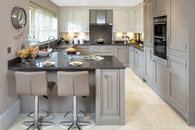 This is an example of a medium sized classic u-shaped kitchen in London with porcelain flooring, beige floors, a double-bowl sink, recessed-panel cabinets, grey cabinets, grey splashback, stainless steel appliances, a breakfast bar and black worktops.