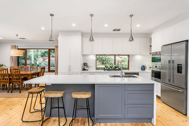 This is an example of a large classic galley kitchen/diner with a double-bowl sink, shaker cabinets, white cabinets, engineered stone countertops, window splashback, stainless steel appliances, light hardwood flooring, an island, brown floors and white worktops.