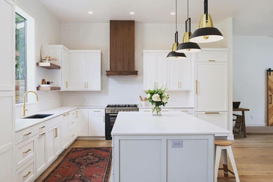 Example of a large trendy l-shaped light wood floor and brown floor kitchen design in Austin with an undermount sink, shaker cabinets, white cabinets, solid surface countertops, white backsplash, subway tile backsplash, paneled appliances, an island and white countertops