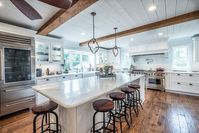 Example of a huge country light wood floor and brown floor eat-in kitchen design in Charlotte with a farmhouse sink, glass-front cabinets, white cabinets, quartz countertops, white backsplash, subway tile backsplash, stainless steel appliances, an island and white countertops