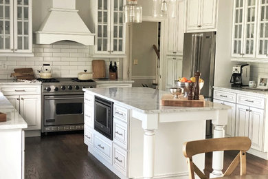 Example of a cottage dark wood floor and brown floor kitchen design in New York with a farmhouse sink, glass-front cabinets, white cabinets, marble countertops, white backsplash, subway tile backsplash, stainless steel appliances, an island and gray countertops