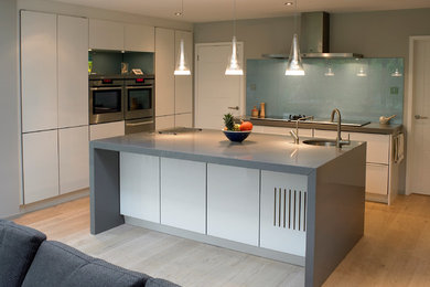 Contemporary grey and white open plan kitchen in Other with a submerged sink, flat-panel cabinets, white cabinets, composite countertops, blue splashback, glass sheet splashback, stainless steel appliances, an island and light hardwood flooring.