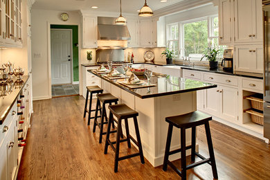 Inspiration for a large timeless u-shaped medium tone wood floor and brown floor enclosed kitchen remodel in New York with a farmhouse sink, shaker cabinets, white cabinets, granite countertops, white backsplash, subway tile backsplash, stainless steel appliances and an island