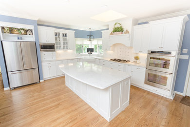 Mid-sized elegant l-shaped light wood floor and beige floor eat-in kitchen photo in Other with a farmhouse sink, recessed-panel cabinets, white cabinets, marble countertops, white backsplash, ceramic backsplash, stainless steel appliances and an island