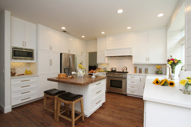 Photo of a large traditional u-shaped kitchen/diner with a belfast sink, recessed-panel cabinets, white cabinets, quartz worktops, white splashback, ceramic splashback, stainless steel appliances, medium hardwood flooring, an island, brown floors and white worktops.