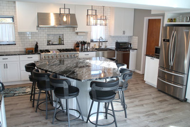 Example of a mid-sized beach style l-shaped vinyl floor and gray floor eat-in kitchen design in Detroit with a farmhouse sink, shaker cabinets, white cabinets, granite countertops, white backsplash, ceramic backsplash, stainless steel appliances, an island and black countertops