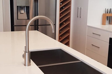 This is an example of a medium sized modern galley kitchen/diner in Adelaide with a submerged sink, flat-panel cabinets, white cabinets, engineered stone countertops, grey splashback, glass sheet splashback, stainless steel appliances, laminate floors, an island and white worktops.