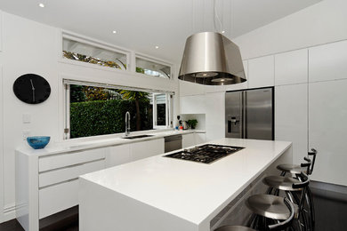 Inspiration for a medium sized contemporary l-shaped kitchen/diner in Auckland with a submerged sink, flat-panel cabinets, white cabinets, composite countertops, stainless steel appliances, dark hardwood flooring, an island and white splashback.