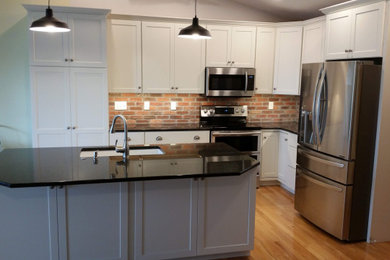 Photo of a medium sized traditional l-shaped kitchen/diner in Wichita with a single-bowl sink, shaker cabinets, white cabinets, granite worktops, red splashback, brick splashback, stainless steel appliances, light hardwood flooring, an island, yellow floors and black worktops.