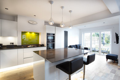 Offene Moderne Küche in L-Form mit flächenbündigen Schrankfronten, weißen Schränken, Küchenrückwand in Grün, Glasrückwand, braunem Holzboden und Halbinsel in Manchester
