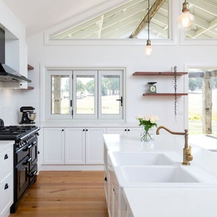 This is an example of a large traditional l-shaped open plan kitchen in Other with a farmhouse sink, recessed-panel cabinets, white cabinets, white splashback, ceramic splashback, black appliances, medium hardwood floors, multiple islands, brown floor, white benchtop and quartz benchtops.
