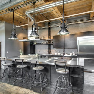 This is an example of a mid-sized industrial galley open plan kitchen in Chicago with a double-bowl sink, flat-panel cabinets, grey cabinets, stainless steel benchtops, grey splashback, stainless steel appliances, light hardwood floors and with island.