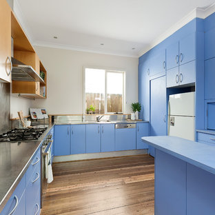 Mid-sized beach style u-shaped separate kitchen in Sydney with an integrated sink, flat-panel cabinets, blue cabinets, stainless steel benchtops, metallic splashback, metal splashback, white appliances, dark hardwood floors, a peninsula and brown floor.