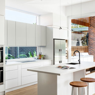 Photo of a contemporary galley open plan kitchen in Sydney with an undermount sink, flat-panel cabinets, white cabinets, window splashback, stainless steel appliances, medium hardwood floors, with island, brown floor and white benchtop.