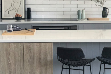 Example of a large trendy galley concrete floor and gray floor open concept kitchen design in Adelaide with an undermount sink, recessed-panel cabinets, gray cabinets, solid surface countertops, white backsplash, ceramic backsplash, black appliances, an island and white countertops