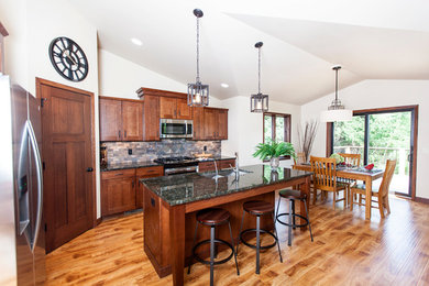 Inspiration for a mid-sized craftsman l-shaped light wood floor eat-in kitchen remodel in Milwaukee with a double-bowl sink, shaker cabinets, dark wood cabinets, granite countertops, multicolored backsplash, stone tile backsplash, stainless steel appliances and an island