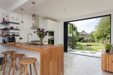 This is an example of a medium sized contemporary l-shaped kitchen/diner in Cheshire with a single-bowl sink, flat-panel cabinets, white cabinets, wood worktops, white splashback, ceramic splashback, stainless steel appliances, travertine flooring, a breakfast bar and grey floors.