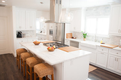 Inspiration for a mid-sized transitional dark wood floor kitchen remodel in Los Angeles with a farmhouse sink, white cabinets, quartzite countertops, white backsplash, stone tile backsplash, stainless steel appliances and an island