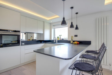 Example of a mid-sized trendy u-shaped open concept kitchen design in London with an integrated sink, flat-panel cabinets, white cabinets, granite countertops, metallic backsplash, glass sheet backsplash, a peninsula and black countertops