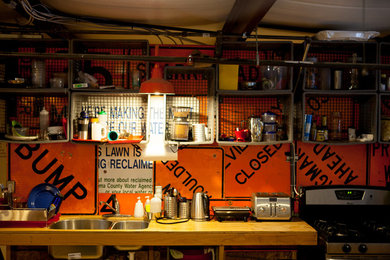 Cette photo montre une petite cuisine américaine parallèle industrielle en bois clair avec un évier encastré, un placard sans porte, un plan de travail en bois, une crédence orange, une crédence en dalle métallique, un électroménager en acier inoxydable, un sol en contreplaqué et aucun îlot.