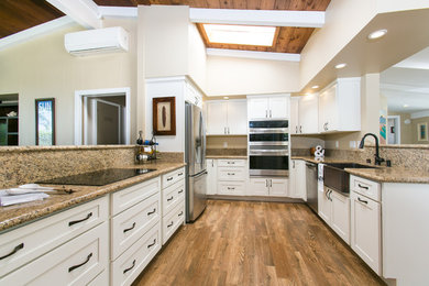 This is an example of a large classic u-shaped open plan kitchen in Hawaii with a belfast sink, shaker cabinets, white cabinets, granite worktops, stainless steel appliances, medium hardwood flooring, an island, brown floors and brown worktops.