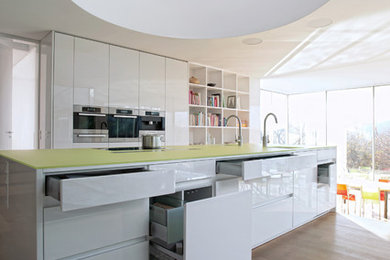 This is an example of a large contemporary open plan kitchen in London with flat-panel cabinets, white cabinets, glass worktops, stainless steel appliances, medium hardwood flooring and an island.