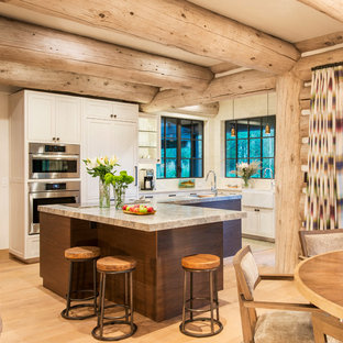 Cette image montre une cuisine américaine chalet en L avec un évier de ferme, un placard à porte shaker, des portes de placard beiges, une crédence en fenêtre, un électroménager en acier inoxydable et un sol en bois clair.