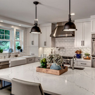 Transitional kitchen inspiration - Example of a transitional l-shaped kitchen design in Seattle with a farmhouse sink, shaker cabinets, white cabinets, white backsplash, subway tile backsplash, stainless steel appliances, an island and white countertops