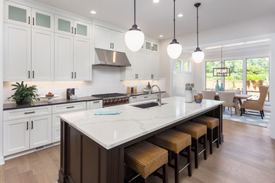 Photo of a large traditional kitchen/diner in San Francisco with a submerged sink, shaker cabinets, white cabinets, quartz worktops, white splashback, ceramic splashback, stainless steel appliances, light hardwood flooring, an island and brown floors.