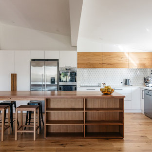Mid-sized beach style l-shaped kitchen in Central Coast with a double-bowl sink, white splashback, ceramic splashback, stainless steel appliances, light hardwood floors, with island, white benchtop, flat-panel cabinets, white cabinets and beige floor.